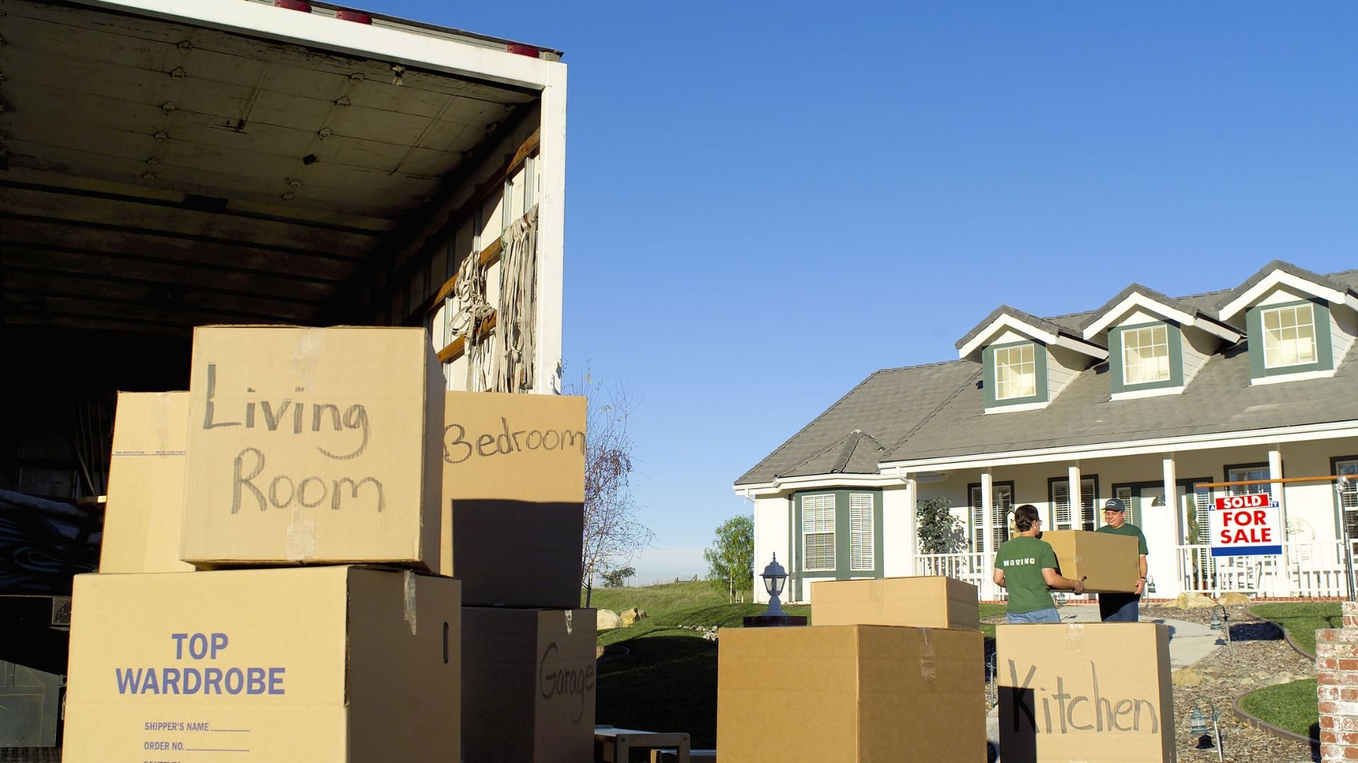 Moving boxes in front of a house with a "Sold" and "For Sale" sign