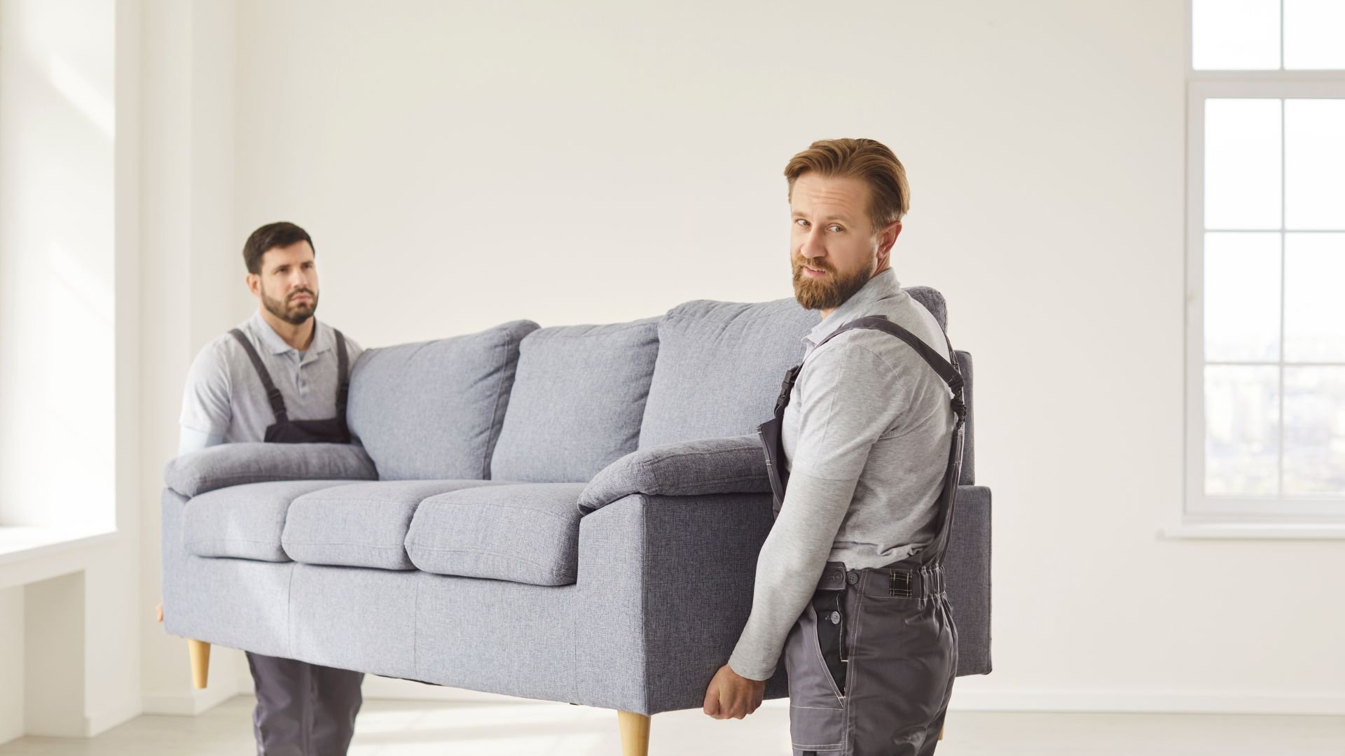 Two movers carrying a gray couch in a bright, empty room